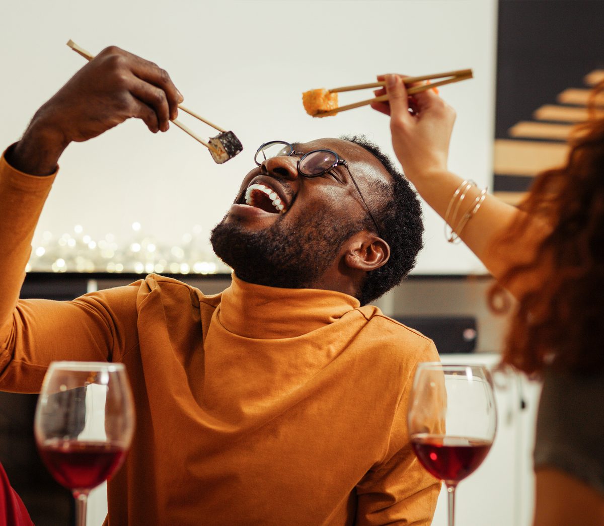 A man with a removable veneer arch in his upper teeth laughs as he lowers a piece of sushi with chopsticks toward his open mouth, a glass of red wine on the table in front of him